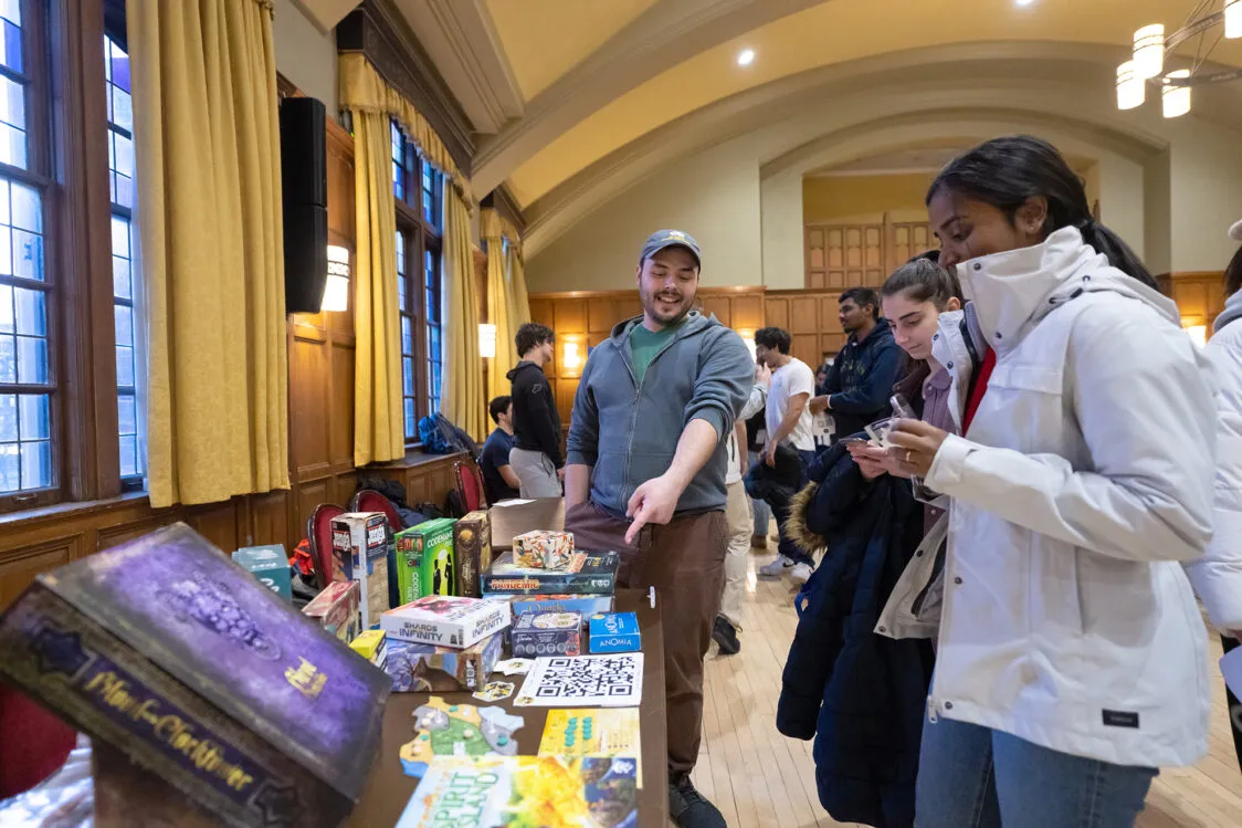 In a wood-paneled hall with tall windows and arched ceilings, students gather around a table of board games and flyers; one person points at items on the display while others look on.