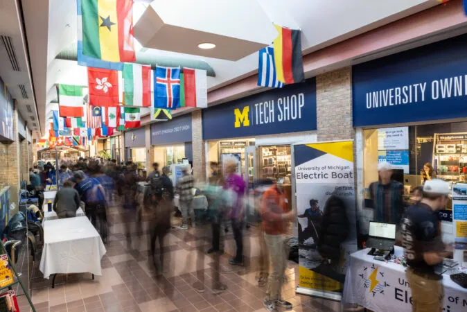 Busy indoor university corridor with international flags hanging overhead; a crowd of students walks past the University of Michigan Tech Shop with tables and a “University of Michigan Electric Boat” display on the right.