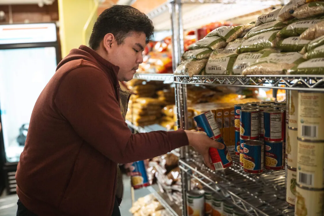 Reza (Ray) Azizan wears a maroon hoodie and is stocking canned goods onto a metal shelf in a food pantry, with bags of rice and other staples stacked above.