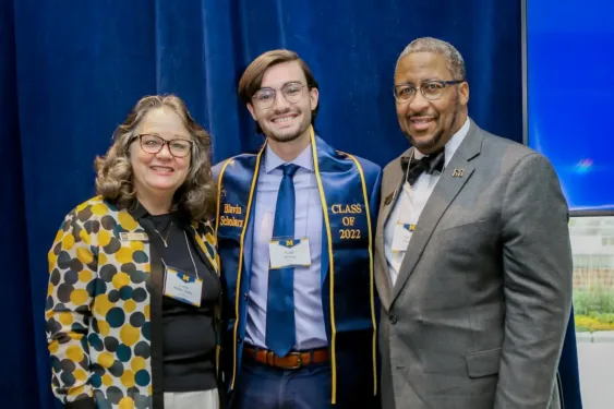 Three people pose for a photo at an event in front of a blue curtain; the person in the center wears a “Blavin Scholars” sash and a name tag.