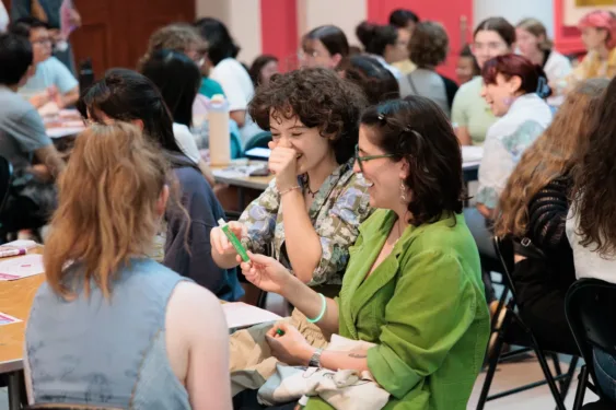 A room filled with students grouped around tables. In the forefront, two women sit at a table together, looking at a book and laughing. One has curly dark hair and is wearing a floral top. The other has dark hair, glasses and a bright green top.