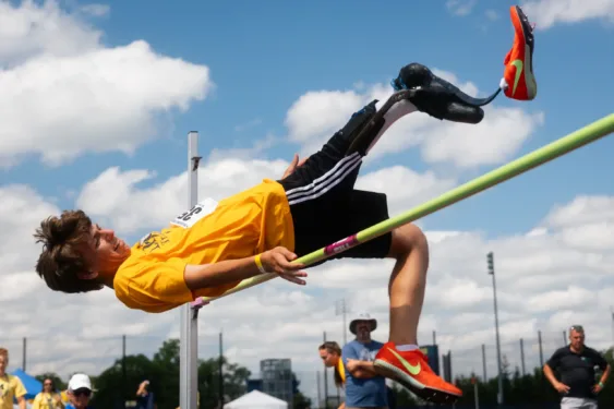 A high jumper with prosthetic leg in a yellow shirt arches backward over the bar during a track-and-field meet, with spectators and a cloudy sky in the background.