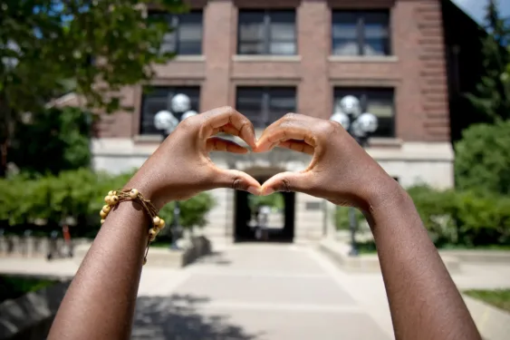 Two hands form a heart shape in the foreground, with a brick campus building and walkway blurred in the background.