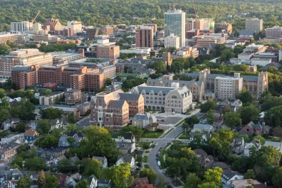 Aerial view of the University of Michigan and surrounding Ann Arbor neighborhoods, showing campus buildings, tree-lined streets, and the city skyline against a wooded hillside.