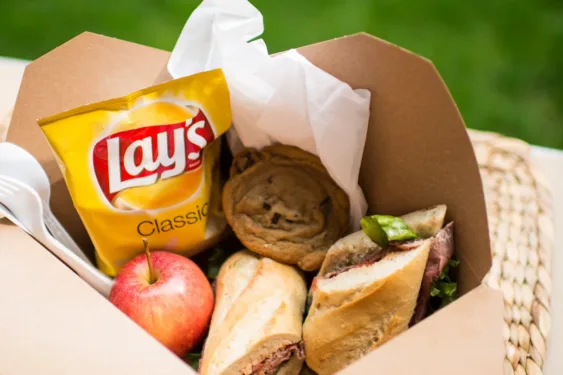 Open cardboard lunch box containing a sandwich, a red apple, a cookie, and a bag of Lay’s Classic potato chips, with napkins and a plastic fork.