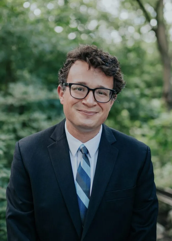 Outdoor head-and-shoulders portrait of a Jacob Lowy in a dark suit jacket, white dress shirt, and blue striped tie, with a softly blurred green leafy background.