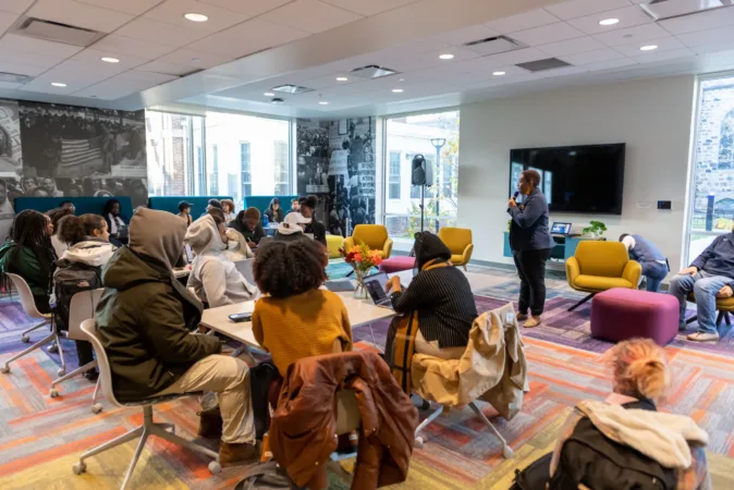 People seated around tables and lounge chairs in a bright meeting room listen to a speaker holding a microphone near a large wall-mounted screen; tall windows and black-and-white photo murals line the walls.