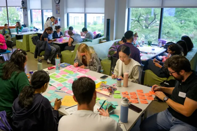 Students gathered around a table working together. They are writing on post it notes.
