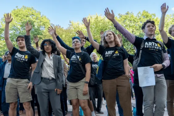 A crowd gathers outdoors with many people raising their hands; several in the front wear black “Arts Resistance” T-shirts, and one person holds a sheet of paper.