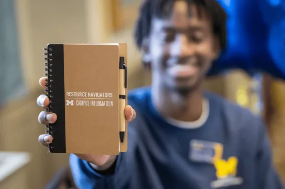 A person holds up a small spiral notebook labeled “Resource Navigators — Campus Information” with a pen attached, while their smiling face is blurred in the background. Button: Visit Campus Information Centers