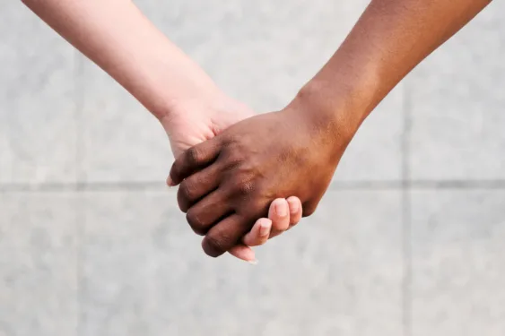 Close-up of two people holding hands, one with a light skin tone and one with a dark skin tone, against a blurred concrete background.