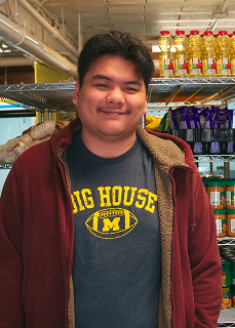 Reza (Ray) Azizan stands in a food pantry wearing a “Big House” Michigan football T-shirt and a maroon hoodie, with shelves of cooking oil and other supplies behind them.