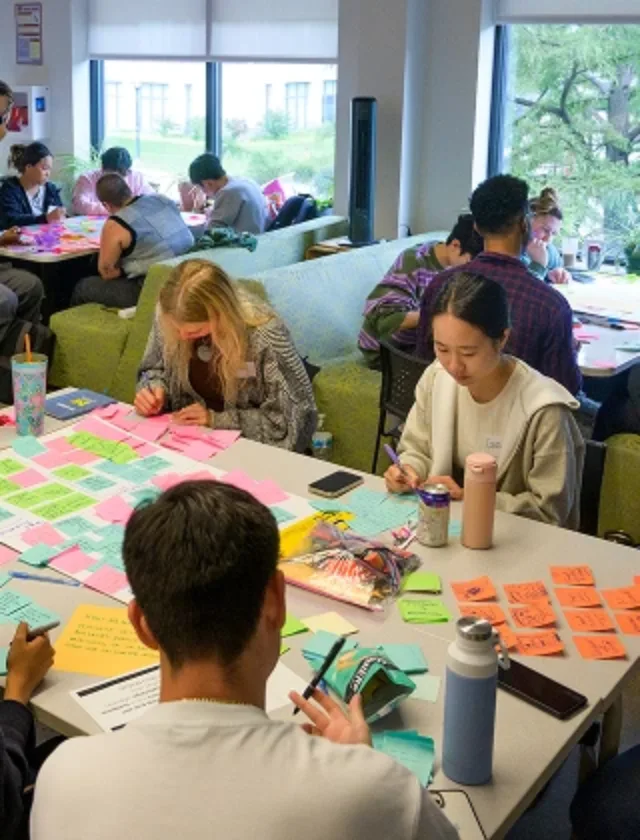 A group of student gathered around a table working together. There are many colorful post it notes stuck to the table.
