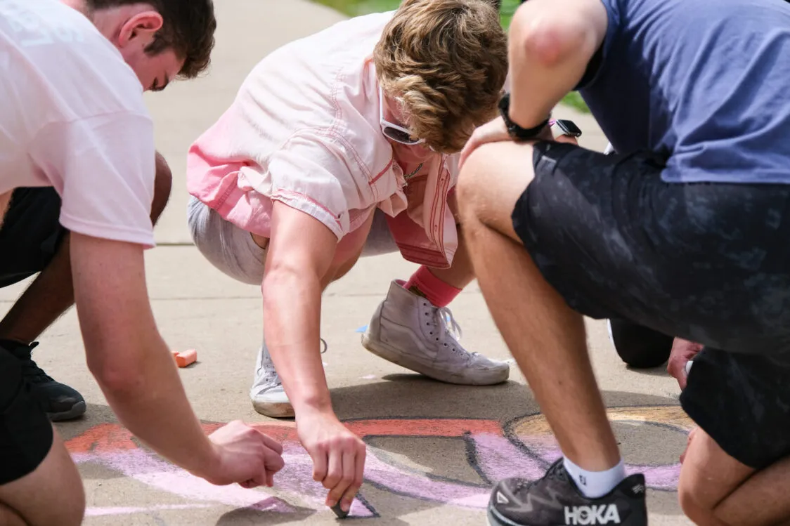 Three people crouch on a sidewalk drawing colorful shapes with sidewalk chalk in bright daylight.