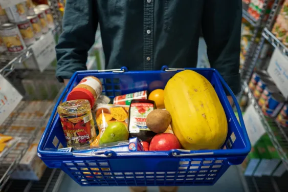 A blue plastic basket of food with fresh fruits and vegetables, and canned goods. The background is slightly blurred and shows wire shelves filled with canned goods.