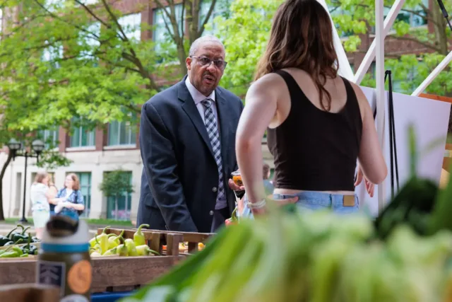 Martino Harmo wears a dark suit and patterned tie and is standing at an outdoor market table holding a small jar, speaking with someone in a black tank top; produce is visible in the foreground.