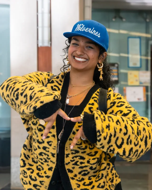 A student wearing a blue baseball cap, gold earrings, and a yellow leopard-print cardigan poses with hands forming an 