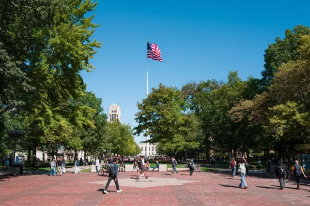 Students walk across the Diag lined with trees and an American flag flying high above with a bell tower visible in the distance under a clear blue sky.