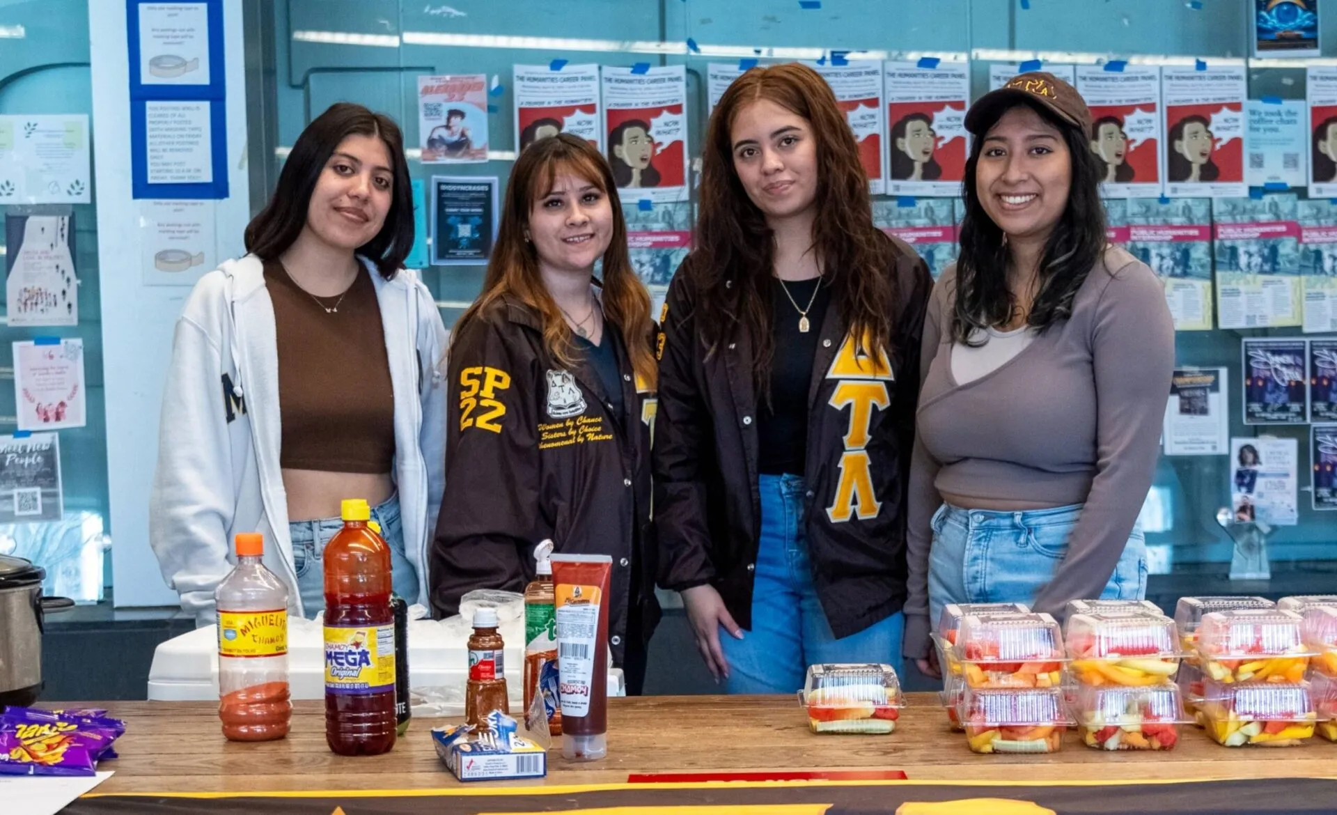 Four students pose behind an information table with a large “ΔΤΛ” banner reading “Delta Tau Lambda Sorority Inc., Alpha Chapter,” with fruit cups, candy, and bottled drinks on the table.
