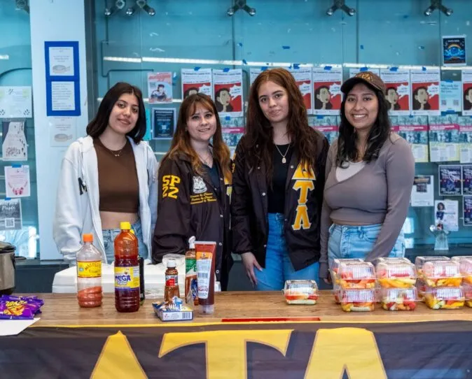 Four students pose behind an information table with a large “ΔΤΛ” banner reading “Delta Tau Lambda Sorority Inc., Alpha Chapter,” with fruit cups, candy, and bottled drinks on the table.