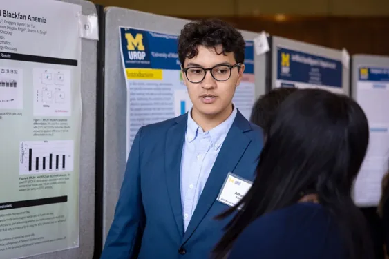 A student in a blue blazer stands beside a University of Michigan UROP research poster display while speaking with another attendee in the foreground; the posters include one titled “Blackfan Anemia” with charts and figures.