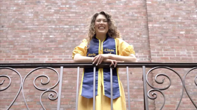 Alana Pollard wears a maize gown and blue stole and is leaning on an ornate wrought-iron railing, with a brick wall filling the background.