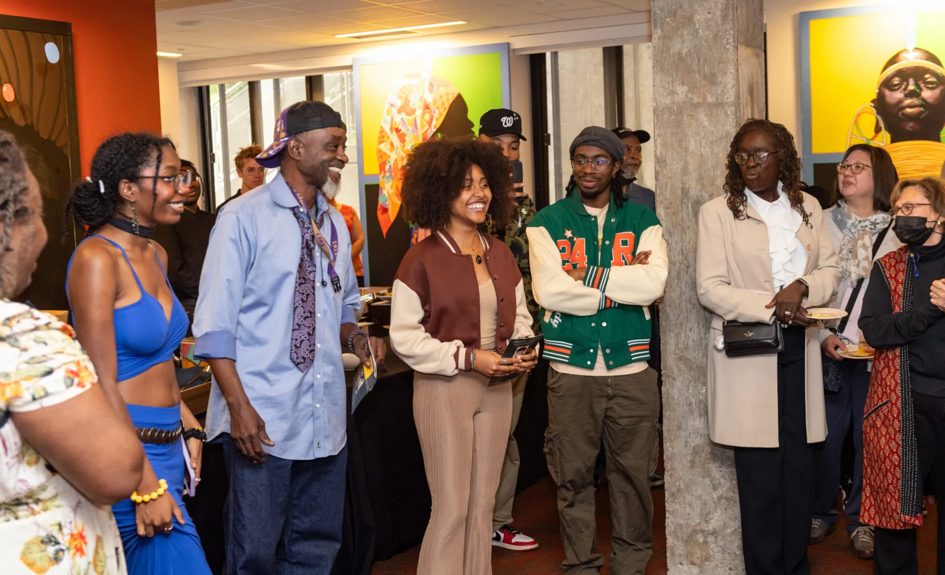 A group of people stands in a brightly lit indoor lobby/gallery space, listening and chatting near large colorful artwork and a concrete pillar; several hold phones or small plates at what appears to be a casual reception or gathering.