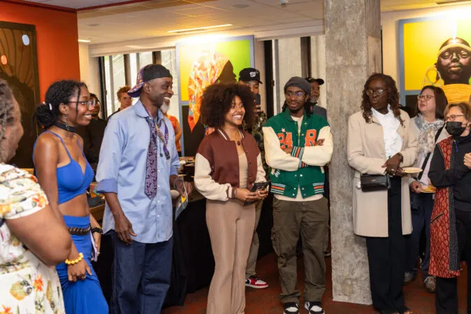 A group of people stands in a brightly lit indoor lobby/gallery space, listening and chatting near large colorful artwork and a concrete pillar; several hold phones or small plates at what appears to be a casual reception or gathering.