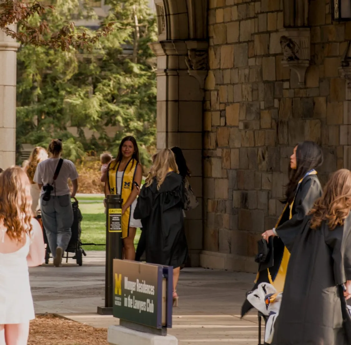 Graduates gather beneath a stone archway on the University of Michigan Ann Arbor campus, with one graduate posing for a photo while others wait nearby.