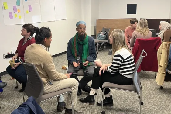 Three students sit in a circle having a discussion. One student has light brown skin and is wearing a hat and scarf. One student has long blonde hair and light skin. One student has light brown skin and dark black hair in a ponytail.