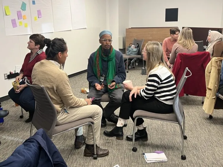 Three students sit in a circle having a discussion. One student has light brown skin and is wearing a hat and scarf. One student has long blonde hair and light skin. One student has light brown skin and dark black hair in a ponytail.