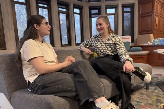 Two students sit on a couch having a discussion. One student has long blonde hair and is smiling as they listen. The other student has dark black hair and is speaking.