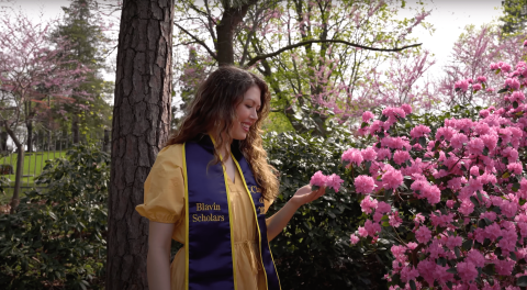 Young blonde woman wearing light yellow dress with a blue sash over shoulders with the text Blavin Scholars printed on it. She is standing in front of trees and admiring pink blossoms.
