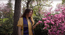 Young blonde woman wearing light yellow dress with a blue sash over shoulders with the text Blavin Scholars printed on it. She is standing in front of trees and admiring pink blossoms.