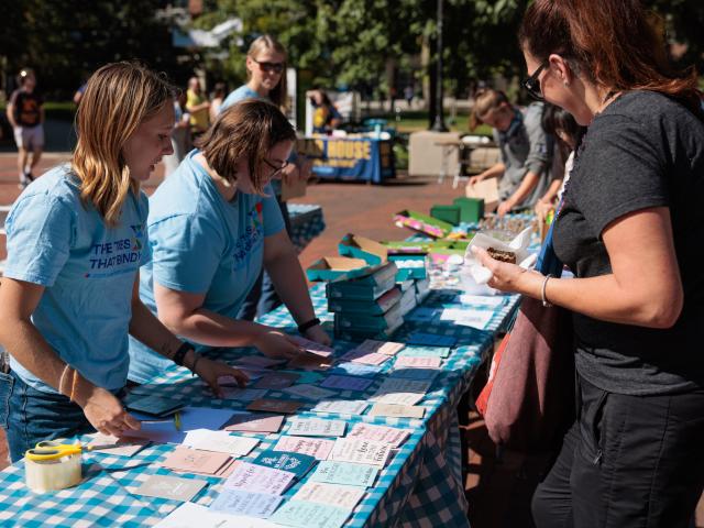 Two young women in light blue shirts stand on one side of a long, rectangular table filled with small notecards. Another woman stands on the opposite side of the table, looking down to examine the table's contents.